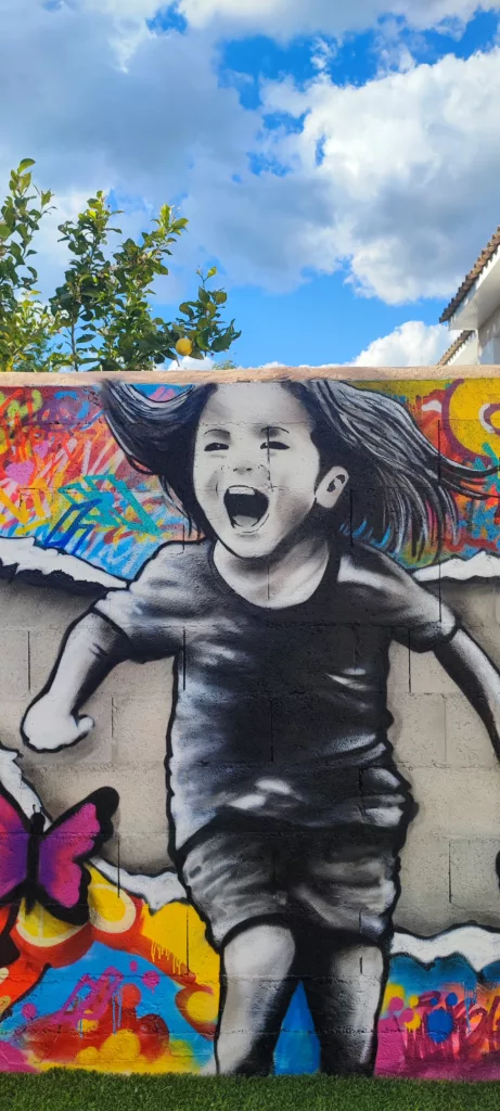 ette fresque graffiti montre deux enfants en noir et blanc courant à travers un mur déchiré, dévoilant un monde coloré rempli de papillons vibrants. Les teintes éclatantes de l’arrière-plan, mêlées à l’inscription « Vivre le bonheur », apportent une énergie positive, célébrant l’enfance, la joie et la liberté